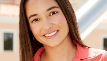 A young woman with straight brown hair smiles at the camera. She is wearing a coral-colored top, and the background is softly blurred with light-colored buildings and windows.