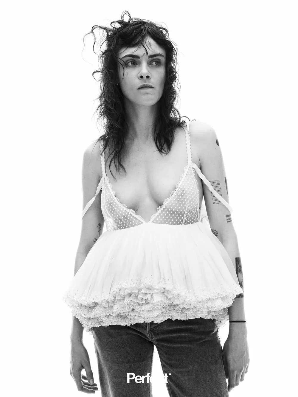 Cara-Delevingne-See-Through-Perfect-Mag1 Black-and-white photo of Cara Delevingne with wavy hair wearing a sheer, ruffled white top and jeans, standing against a plain background with a serious expression. The word Perfect appears at the bottom of the image.