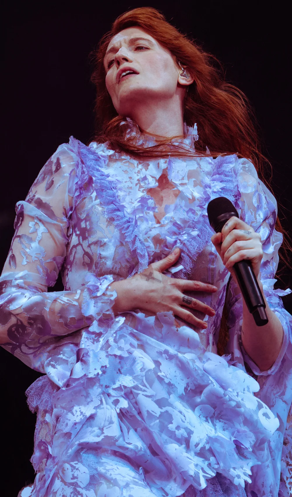 Florence-Welch-2026-Leeds-Festival A performer in a sheer, ruffled, light purple dress sings passionately on stage, holding a microphone in one hand and clutching their chest with the other.
