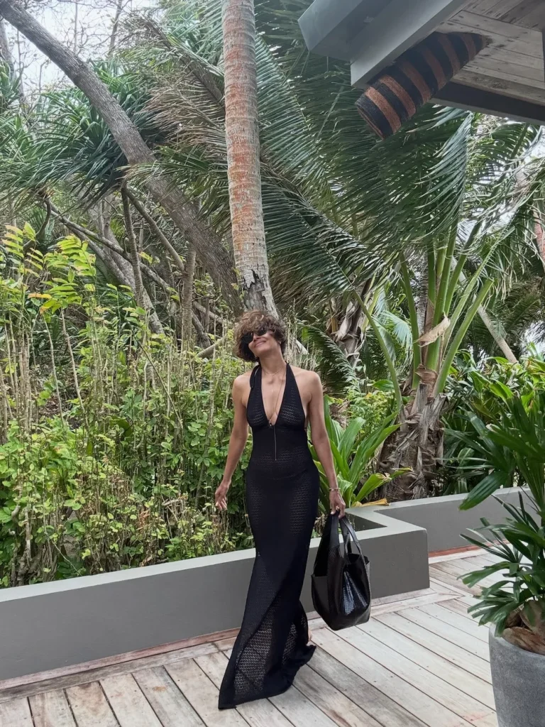 Halle Berry in a sleeveless black dress and sunglasses stands on a wooden deck surrounded by lush green tropical plants and palm trees, holding a large black bag.