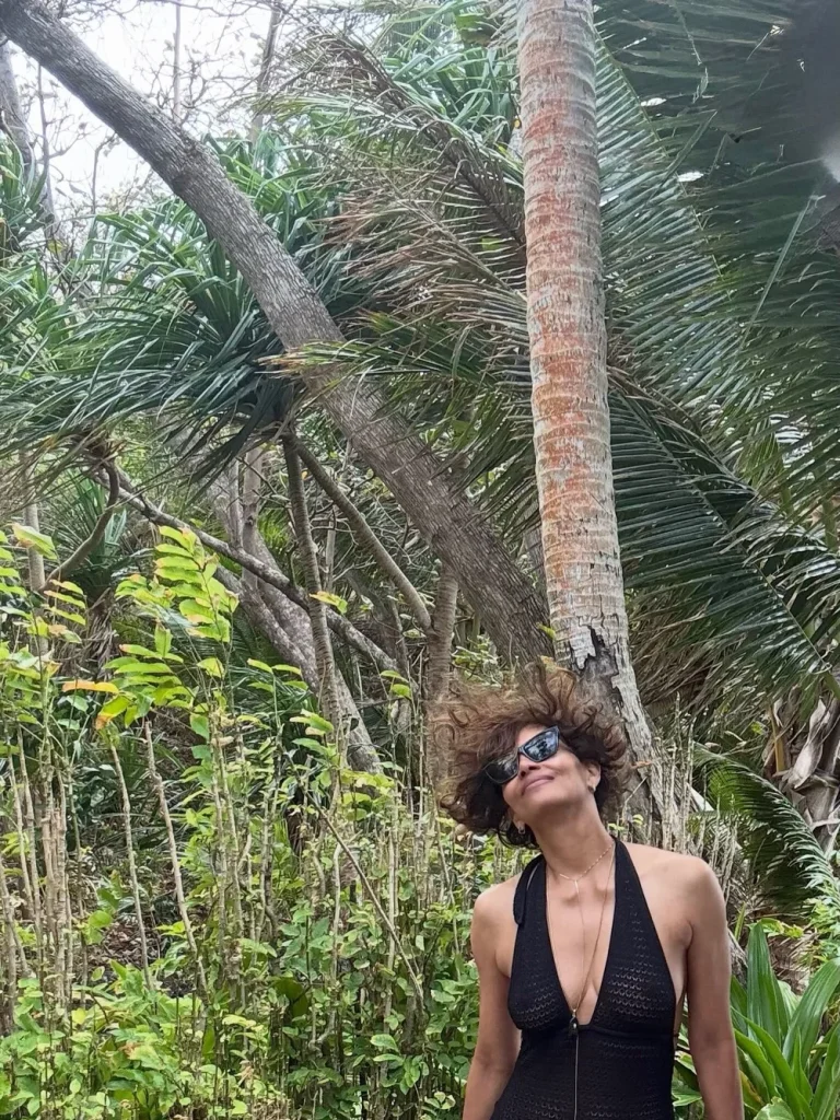 Halle Berry wearing sunglasses and a black swimsuit stands in front of tall, wind-blown palm trees and lush greenery, looking up with hair blown to the side.