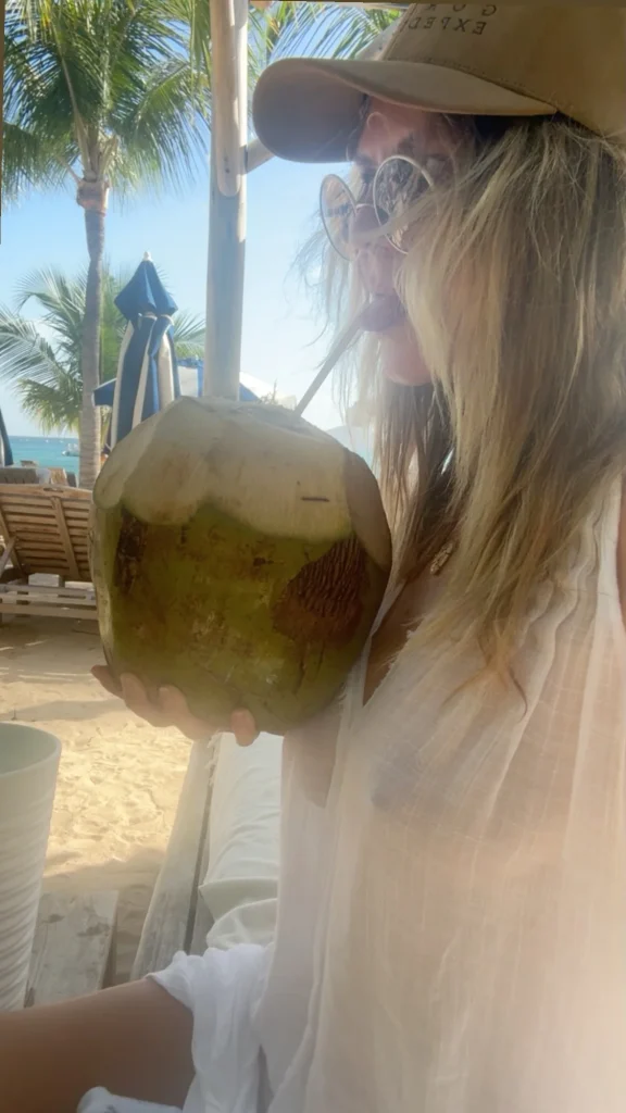 A woman in sunglasses and a hat drinks from a large coconut with a straw on a sunny beach, with palm trees, lounge chairs, and umbrellas visible in the background.