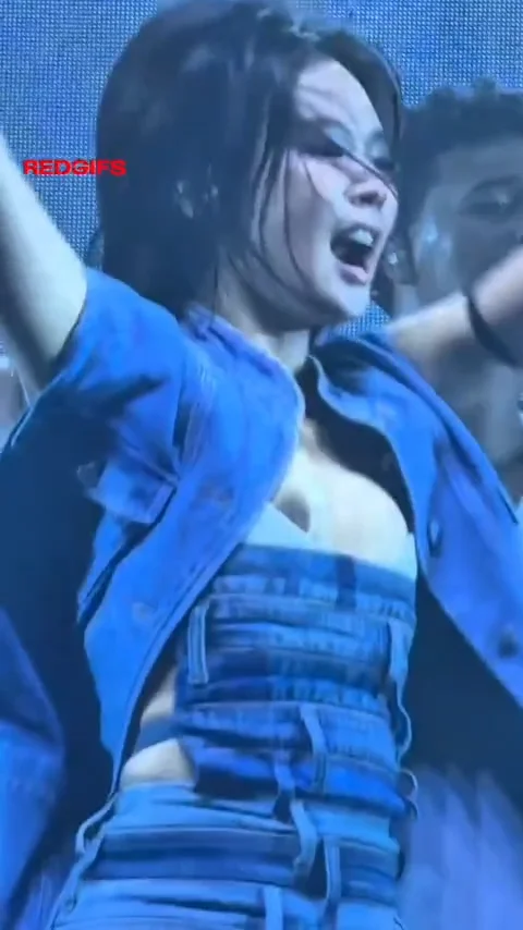 A woman in a blue denim outfit performs energetically on stage, arms raised, with other dancers visible in the background and a large LED screen behind her.