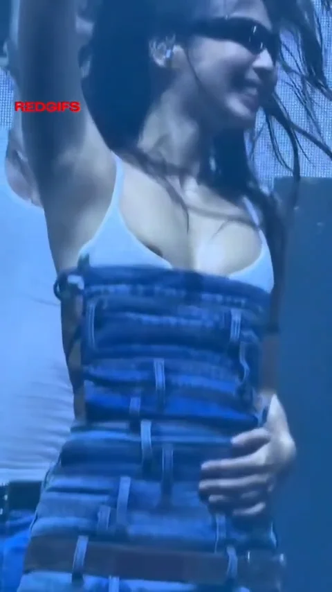 A woman in a stylish dress and sunglasses performs on stage, smiling with her arm raised. Blue lighting creates a vibrant atmosphere.