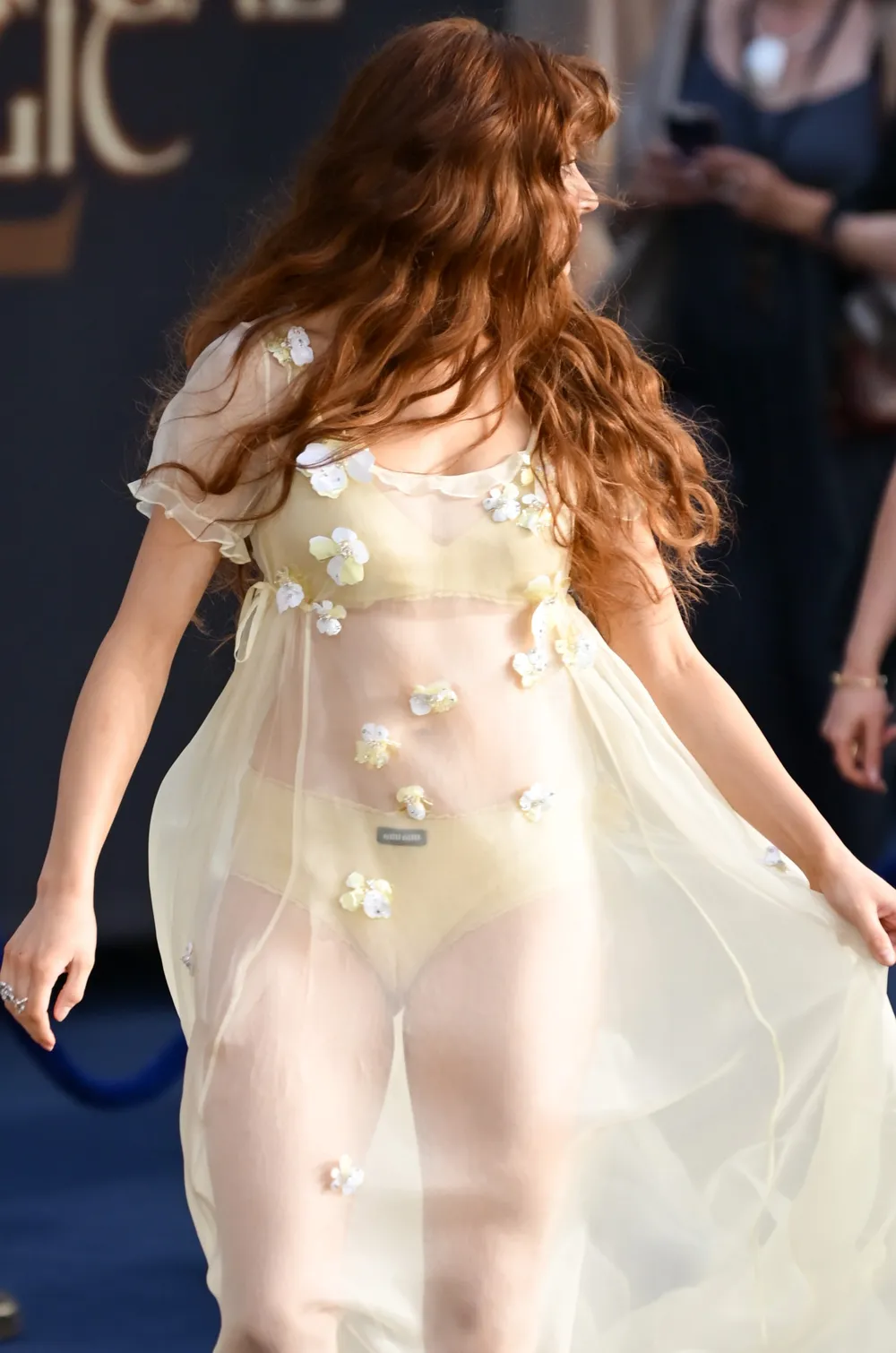 Joey-King-Practical-Magic-2-Camel-Toe A woman with long red hair wears a sheer, pale yellow dress with floral appliqués over matching underwear, walking outdoors at an event.