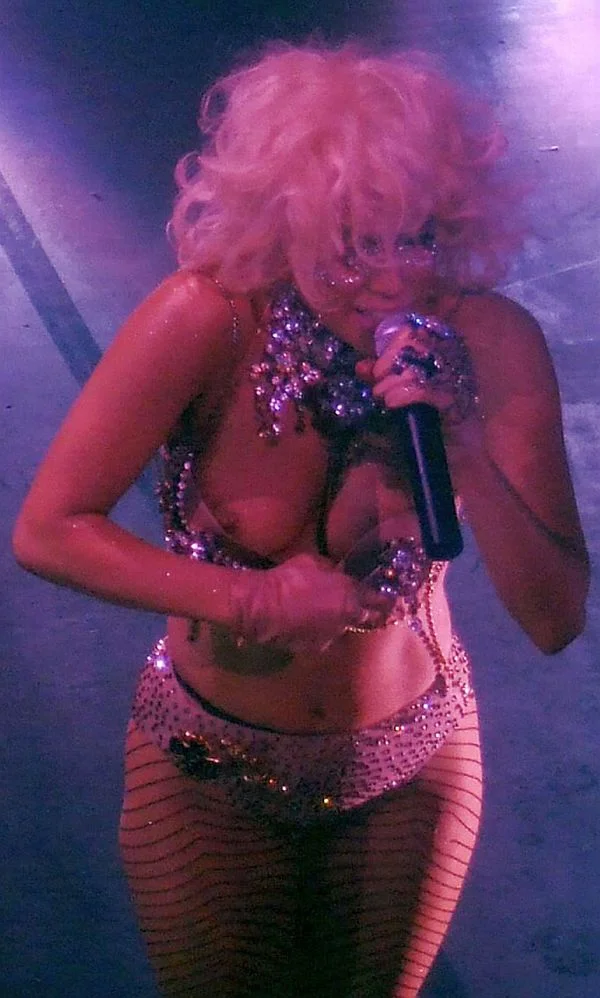 Lady Gaga with platinum blonde curly hair performs on stage, wearing a sparkling jeweled outfit, fishnet stockings, and holding a microphone, under purple lighting.