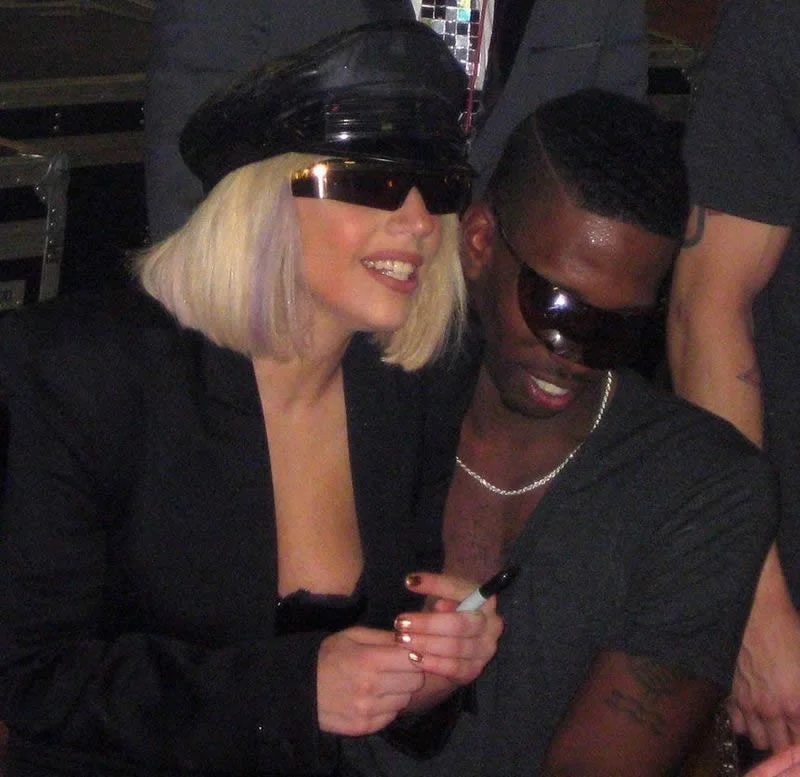 Lady Gaga and a man sit closely together, both wearing dark sunglasses. Lady Gaga has short blonde hair and a black hat, smiling while holding a marker. The man has short dark hair and is wearing a grey shirt.