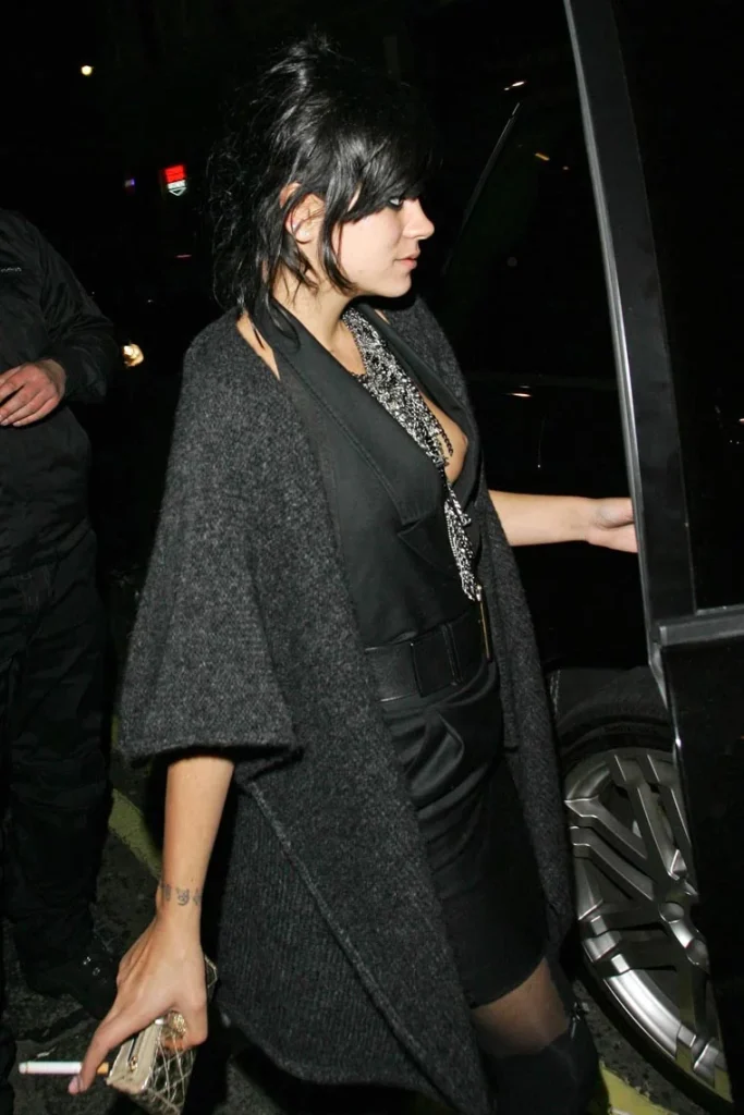 A woman with dark hair in a ponytail, wearing a black dress, gray cardigan, and necklace, holds a cigarette and a small clutch while stepping into a car at night.