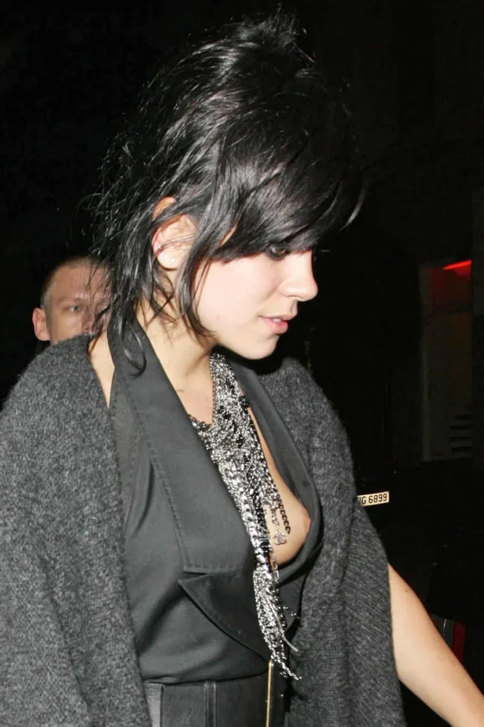 A woman with dark hair wearing a black outfit and layered silver necklaces looks down while walking at night, with another person partially visible in the background.