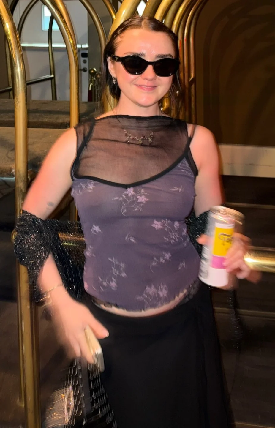 A woman wearing black sunglasses, a sheer black top over a floral tank, and a black skirt holds a can and a small bag. They are standing indoors in front of gold luggage carts.