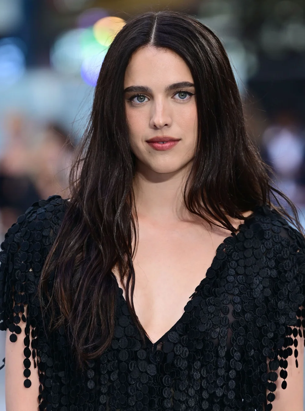 A woman with long dark hair and blue eyes wears a black, textured, deep V-neck dress. She stands outdoors, looking at the camera with a neutral expression. The background is blurred and features colorful lights.
