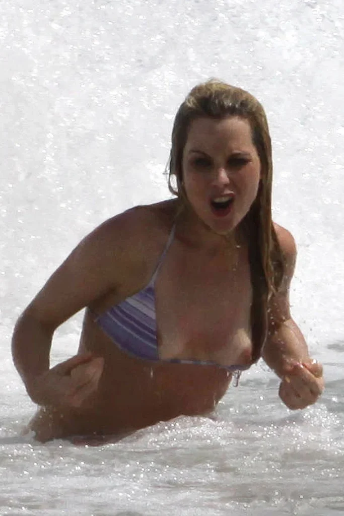 A woman in a striped bikini stands in shallow ocean water, appearing to shout or make an expressive face, with water splashing around her.
