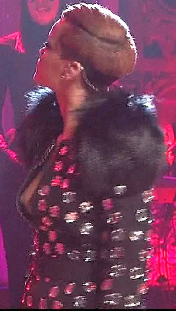 A person with short, styled hair wearing a black outfit decorated with shiny spots and large black fur shoulders, standing on a stage with pink lighting and another person blurred in the background.