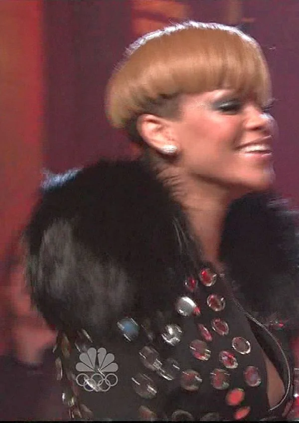 Smiling woman with short, light brown hair and bangs, wearing a black jacket with shiny studs and a fur collar, standing in front of a blurred red background.