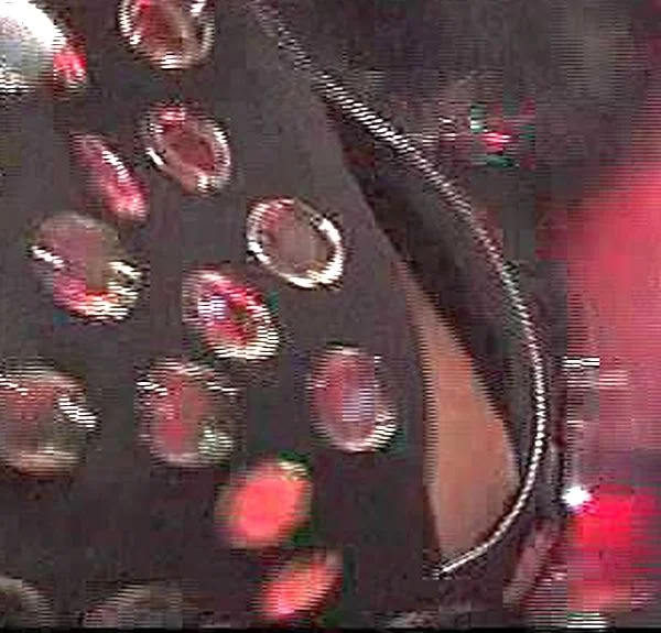 A close-up of black fabric with shiny metallic circular cutouts, reflecting red and white lights, possibly part of a costume or clothing, with a blurred and colorful background.