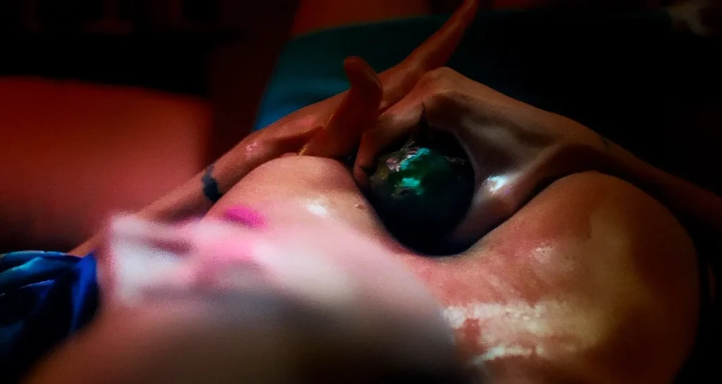 A dark, smooth massage stone being held against oiled skin between the breasts of a woman. Its a close up, viewed from above. The partial face of the woman is blurry. Photo 1 / 4.