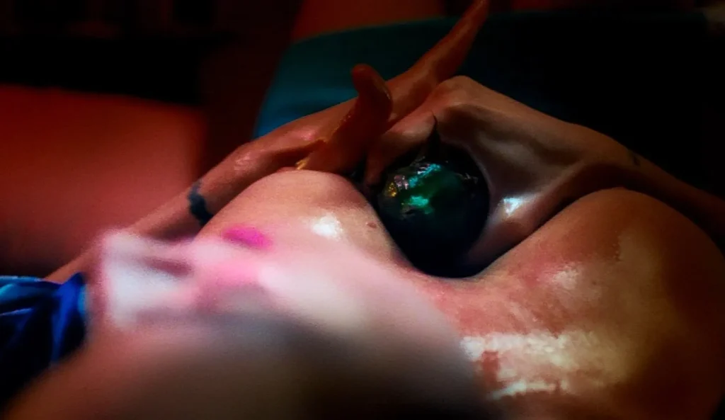 A dark, smooth massage stone being held against oiled skin between the breasts of a woman. Its a close up, viewed from above. The partial face of the woman is blurry. Photo 3 / 4.