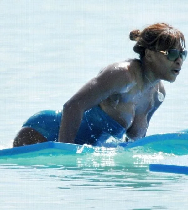 Woman in a blue swimsuit and sunglasses, wet from the water, leaning forward on a blue surfboard or paddleboard in a calm, light blue sea.