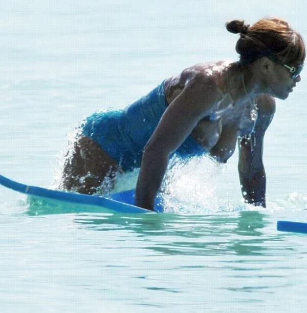 A woman in a blue swimsuit and sunglasses is kneeling on a blue surfboard in clear, shallow water.