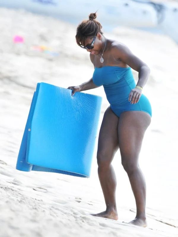 A woman in a blue swimsuit holds a blue mat while standing on a sandy beach. She is wearing sunglasses and a necklace, with her hair tied up.