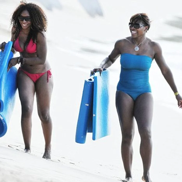 Two women in swimsuits walk on a sandy beach, each holding a blue foam mat. One wears a red bikini, the other a blue one-piece. Both are smiling and enjoying their time by the sea.