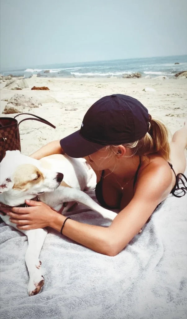 Woman in a black cap and swimsuit lying on a beach towel, hugging a white and brown dog. The beach and ocean are in the background.