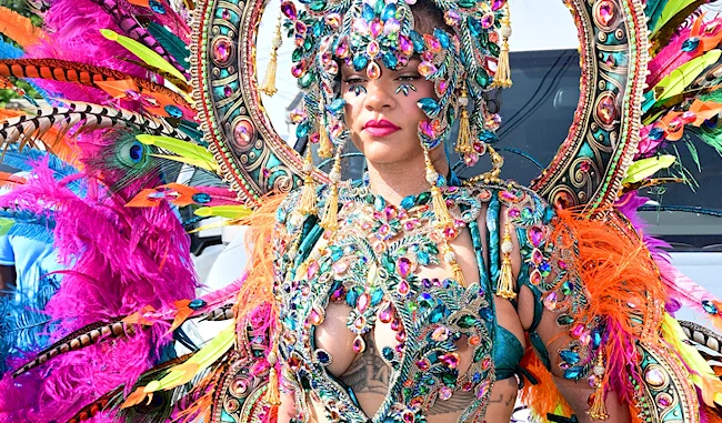 Rihanna wearing an elaborate, colorful costume with feathers, beads, and jewels at a festival or parade, with vibrant pink, orange, blue, and green accents.
