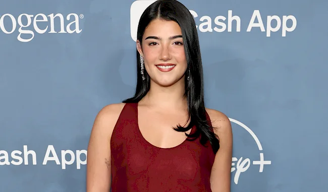 A woman with long dark hair in a sleeveless burgundy dress smiles at a blue step-and-repeat event backdrop featuring company logos.
