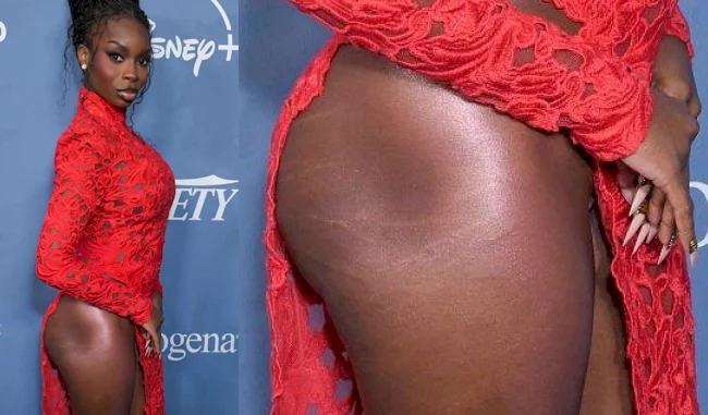 A black woman in a red lace dress poses on a blue carpet with a side view, highlighting a close-up of their exposed hip and thigh.