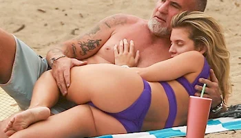 A shirtless man with tattoos lies on the sand, holding a woman in a purple bikini as she rests her head on his chest. They are relaxing on a beach blanket with a pink tumbler nearby.