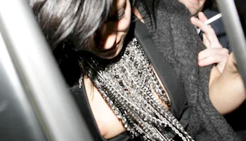 A woman with dark hair and a sparkling necklace leans forward in a dimly lit space while another person holds a cigarette in the background.