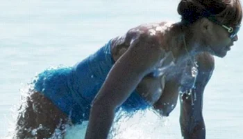 Serena Williams in a blue swimsuit is emerging from the water, wearing sunglasses. The background is light, suggesting daylight and a beach or pool setting.