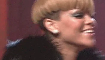 Rihanna with short, light brown hair and earrings is smiling, wearing a dark fur-like garment, with a blurred red background.