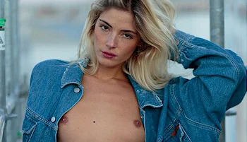 A fair-skinned woman with blonde hair, styled in a relaxed, messy fashion, wears an unbuttoned blue denim jacket. She has a natural look, looking directly at the camera with a neutral expression. The background appears to be outdoors.