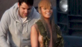 Rihanna smiling and looking away from the camera stands next to a man in a gray hoodie; they are outdoors, and a large vehicle is in the background.