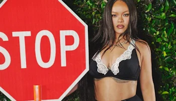 A woman in a black and white lace outfit stands next to a large red stop sign, with green foliage in the background.