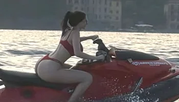 A woman in a red bikini rides a red jet ski on the water, with buildings and trees visible in the background during sunset.