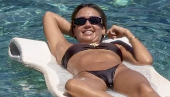 A woman wearing sunglasses and a dark bikini relaxes on a float in a pool, smiling with her arms behind her head.
