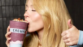 A woman with long blonde hair kisses a large purple smoothie topped with granola, labeled EREWHO, while giving a thumbs up.