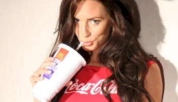 A woman with long brown hair wearing a red Coca-Cola shirt drinks from a large McDonalds cup using a straw.