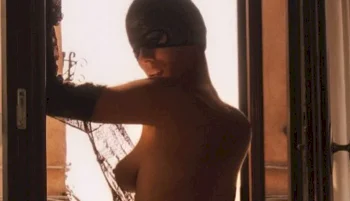 A masked, topless woman stands in a sunlit doorway, reaching toward broken glass. Dramatic lighting creates a silhouette effect against warm, golden tones.