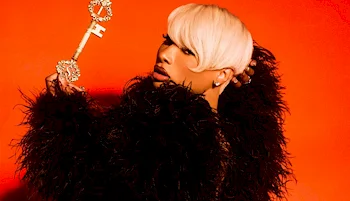 A woman with short blond hair in a black feathered outfit holds a large, ornate gold key against a bright orange background.