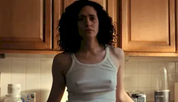 A woman with curly dark hair stands in a kitchen, wearing a white tank top. Wooden cabinets and kitchen items are visible in the background. The lighting is dim and her expression appears serious.