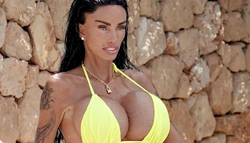 A woman with long dark hair, wearing a bright yellow bikini, stands in front of a stone wall. She has visible tattoos on her arm and is looking slightly to the side.