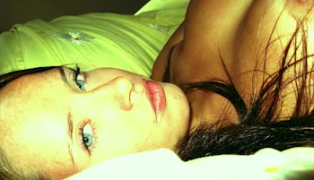 A woman with long dark hair and blue eyes lies on a green pillow, looking at the camera. The lighting is bright and warm, highlighting their facial features.