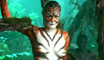 A woman with body paint resembling a tiger stands in a jungle setting, with green foliage and a branch covered in moss. Another figure is partially visible in the background.