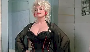 A woman with curly blonde hair and red lipstick wears a black corset with red trim and a sheer black robe, standing confidently with her hands on her hips in a tiled room.