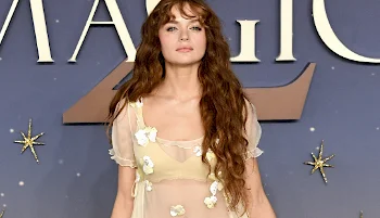 A woman with long wavy hair in a sheer, floral-embellished dress poses in front of a blue background with large gold letters and star decorations.