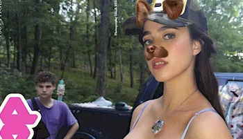 A young woman with a dog filter on her face stands outdoors, wearing a cap and necklace. In the background, a boy in a purple shirt sits near a car in a wooded area.