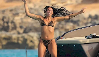 A woman in a leopard-print bikini stands on a boat with arms raised, smiling, with rocky cliffs and water in the background.